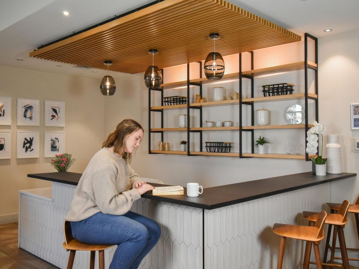 The Vicinity woman reading a book at a modern kitchen counter with shelves and hanging lights in the background.