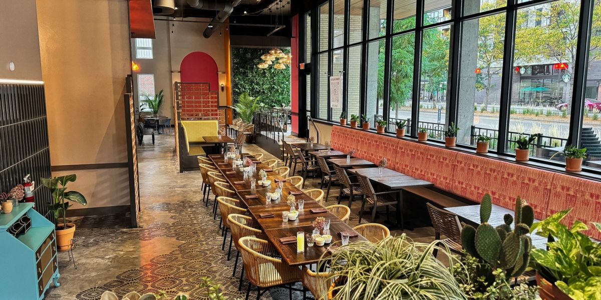 A trendy bar area at La Madre Mexican restaurant in The Vicinity apartments, featuring ample greenery, colorful furnishings, large windows, and a variety of seating options.